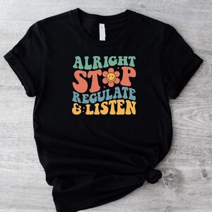 May include: Black t-shirt featuring the phrase "ALRIGHT STOP REGULATE & LISTEN" in a retro font, with a smiling flower graphic. The text is in orange, green, blue, and yellow. A casual, everyday garment.