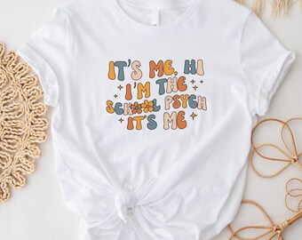 May include: White t-shirt with a colourful graphic that says "It's me, hi I'm the school psych it's me".