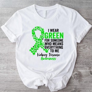 Puede incluir: Camiseta blanca con un diseño de lazo verde hecho de hojas y el texto: "Llevo verde por alguien que lo es todo para mí. Concienciación sobre la enfermedad renal."