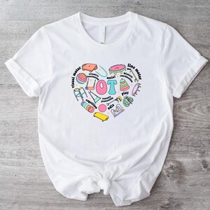 May include: White t-shirt with a pink heart design featuring the word "OT" surrounded by various colourful illustrations of objects related to occupational therapy, including a walking frame, a ball, a skateboard, and a drum. The text around the heart includes "visual motor", "fine motor", "play", "cognition", "sensory processing", "social interaction", "executive functioning", and "ADL's".