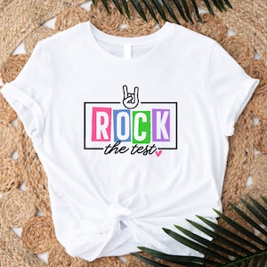 May include: White t-shirt with a colorful graphic that says "ROCK the test" with a pink heart and a hand making a rock and roll sign.