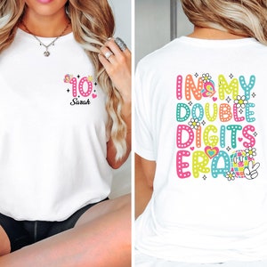 Könnte beinhalten: Weißes T-Shirt mit einer bunten Grafik auf der Rückseite, die "In My Double Digits Era" mit einem Peace-Zeichen und einer Discokugel zeigt. Die Vorderseite des Shirts hat eine rosa Zahl 10 mit Sternen und den Namen "Sarah" darunter.