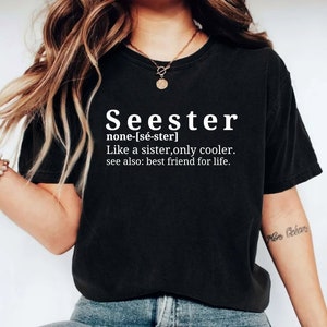 以下が含まれることがあります： 黒いTシャツに白い文字で「Seester」と書いてあり、その単語の定義が書かれています。定義は「none-[sé-ster] 姉妹のような存在、でももっとクール。別名：生涯の親友。」です。