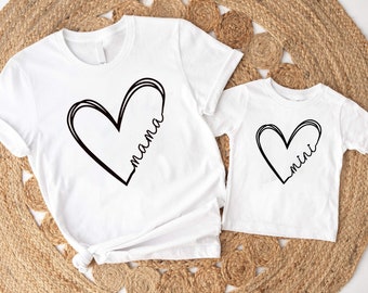 Könnte beinhalten: Zwei weiße T-Shirts mit schwarzen Herzmotiven. Das größere Shirt sagt "mama" im Herzen und das kleinere Shirt sagt "mini" im Herzen.