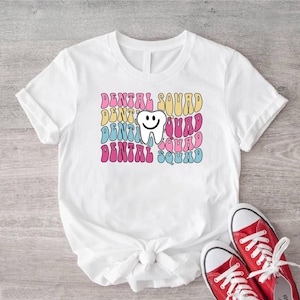 Könnte beinhalten: Weißes T-Shirt mit dem Text "DENTAL SQUAD" in einem Retro-Stil, gestapelt in Rosa, Gelb, Blau und Rosa. Eine lächelnde Zahn-Grafik ist auf dem Design zentriert. Rote Leinenschuhe mit weißen Schnürsenkeln unten.