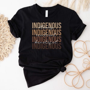 May include: Black t-shirt with the text "INDIGENOUS" repeated five times in brown and beige lettering. The text is stacked vertically with the words "strong", "resilient", and "beautiful" written in cursive between the words "INDIGENOUS".