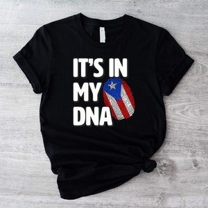 Puede incluir: Camiseta negra con texto blanco que dice "It's in my DNA" y un diseño de huella dactilar con los colores de la bandera puertorriqueña.