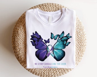 Puede incluir: Camiseta blanca con un diseño de mariposa morada y turquesa. Las mariposas están formadas por mariposas más pequeñas. El texto "NO STORY SHOULD END TOO SOON" y "#suicideawareness" están impresos en la camiseta.