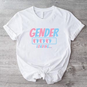 May include: White t-shirt with the text "GENDER LOADING..." in pink and blue. The design includes baby footprints in pink and blue, suggesting a gender reveal theme. The shirt is knotted at the waist.