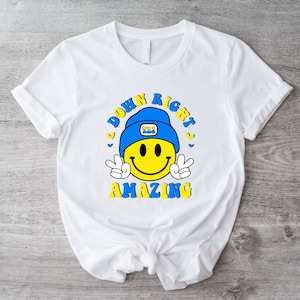 Op de afbeelding: Wit T-shirt met een gele smiley die een blauwe hoed draagt en de tekst "Down Right Amazing" in blauwe en gele letters.