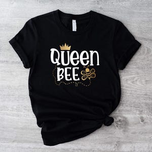 May include: Black t-shirt with a gold crown and the text "Queen Bee" in white. A gold bee with black stripes is next to the text.