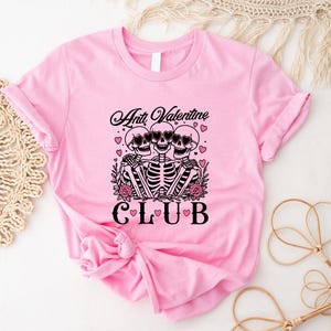 Puede incluir: Camiseta rosa con un gráfico de tres esqueletos con gafas de sol y el texto "Anti Valentine Club".