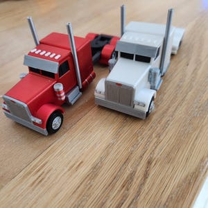 May include: Two toy semi-trucks, one red and one white, featuring silver grilles, tall exhaust stacks, and black wheels. The red truck is positioned slightly behind the white truck, and both models display detailed cab structures with rectangular windows.