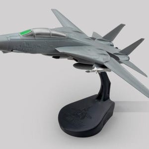 May include: A grey model aeroplane with a green cockpit window, mounted on a black stand. The model has detailed wings and a sleek design, showcasing a miniature aircraft replica. The stand has a logo.