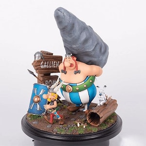 Puede incluir: Figura detallada con personajes de la serie de cómics Astérix. Obélix lleva una gran piedra. Astérix está cerca con su escudo. También hay un perro pequeño y un letrero que dice "Gallien" y "Rom".
