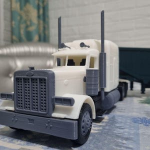 May include: A detailed model of a white and grey articulated lorry with a trailer. The lorry has a prominent front grill, headlights, and exhaust stacks. The trailer is dark green. The model is likely 3D-printed and sits on a patterned surface.