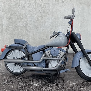May include: A blue and gray toy motorcycle with a detailed engine, exhaust pipes, and fenders. The model has a classic cruiser design with a seat, handlebars, and a headlight. The wheels are white with black tires. The motorcycle is set against a neutral background.