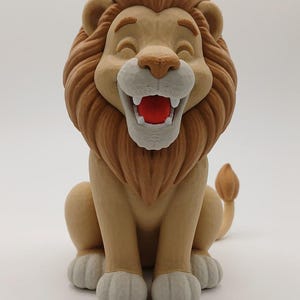 May include: A 3D printed lion figurine in a seated position with a tan body, a darker brown mane, and light gray paws. The lion has a cartoonish expression with a wide, open mouth revealing a bright red interior and white teeth.