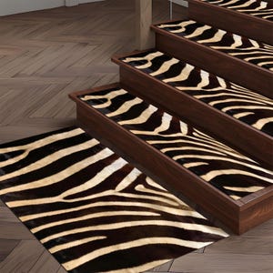 Puede incluir: Peldaños de escalera y una alfombra a juego con un estampado de cebra. Las huellas están instaladas en escaleras de madera marrón oscuro. La alfombra es rectangular y está colocada en el suelo. El estampado de cebra tiene rayas negras y crema alternas.