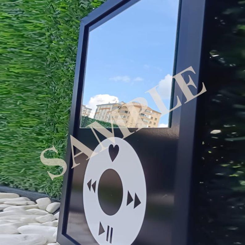 May include: Black framed mirror with a white graphic of a music player's control buttons. The mirror reflects a blue sky with clouds and buildings. The word "SAMPLE" is partially visible.
