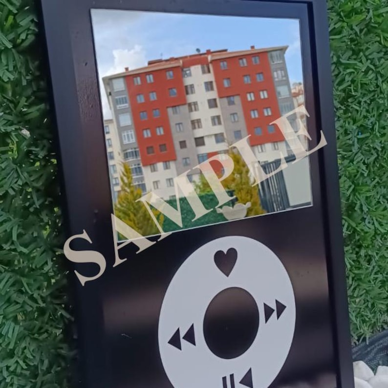 May include: Black framed mirror with a white graphic of a music player's control buttons. The mirror reflects a building with red and white walls and a blue sky. The word "SAMPLE" is printed across the mirror.