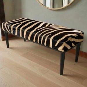 May include: A zebra-print bench with black legs. The seat has a pattern of black and tan stripes. The bench is made of wood and is a stylish piece of furniture. The bench is approximately 120 cm long.