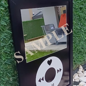May include: Black framed mirror with a white graphic of a music player's control buttons. The mirror reflects an outdoor scene with patio furniture and the word "SAMPLE" overlaid on the mirror's surface.