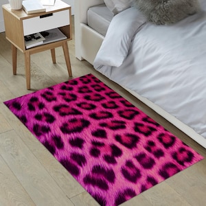 May include: A pink and black leopard print rug with a fuzzy texture. The rug is placed on a wooden floor in front of a white bed.