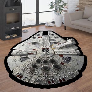 Puede incluir: Una alfombra redonda con un borde negro que muestra una imagen blanca y gris del Millennium Falcon, la nave espacial de Star Wars. La nave espacial se representa con detalle, con puertos de escape de motor visibles y otras características.