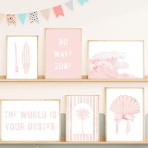 May include: A collection of framed art prints with a beach theme. The prints feature a surfboard, the text "NO WAKE ZONE", a watercolor wave, the text "THE WORLD IS YOUR OYSTER", a palm tree, and a seashell. The color palette is soft pink and white.