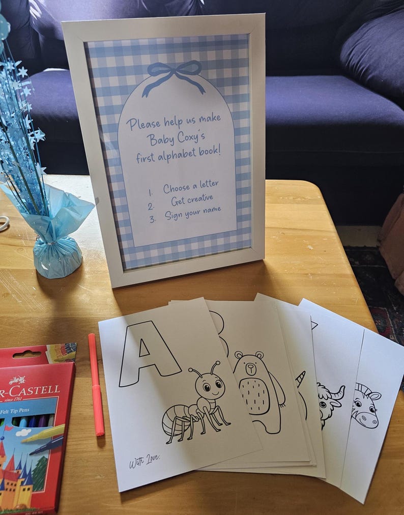 May include: A framed sign with a blue gingham pattern and the text "Please help us make Baby Coxy's first alphabet book!" is displayed. Beside it are coloring pages with the letter "A" and an ant illustration. A box of felt tip pens and a pink marker are also visible.