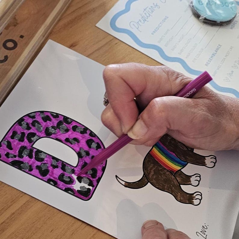 May include: A person coloring a baby shower game. The game includes a drawing of a dog with a rainbow collar and the letter "D" with a pink leopard print design. The word "Predictions" is visible at the top of the page.