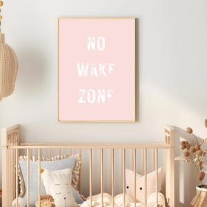 May include: A framed pink print with the words "NO WAKE ZONE" in white, displayed above a wooden crib. The room is decorated with a woven pendant light and decorative pillows. A vase of dried flowers sits nearby.