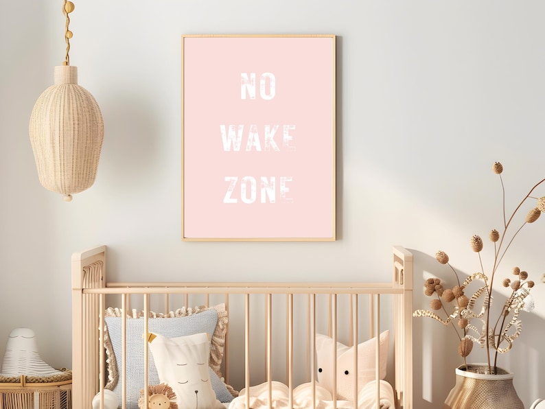 May include: A framed pink print with the words "NO WAKE ZONE" in white, displayed above a wooden crib. The room is decorated with a woven pendant light and decorative pillows. A vase of dried flowers sits nearby.