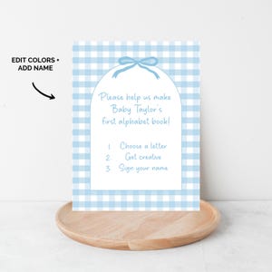 May include: A baby shower invitation with a blue gingham pattern background. The invitation reads "Please help us make Baby Taylor's first alphabet book!" with instructions to choose a letter, get creative, and sign your name.