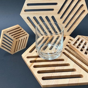 May include: Set of four wooden coasters with a geometric design. The coasters are made of light wood and have a natural finish. A clear glass is sitting on top of the coasters.