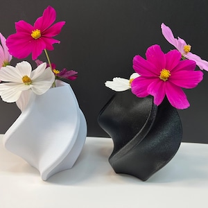 Puede incluir: Dos jarrones decorativos, uno blanco y otro negro, cada uno con coloridas flores de cosmos. Los jarrones tienen un diseño retorcido único. Las flores son rosas, blancas y moradas con centros amarillos. El fondo es gris oscuro.