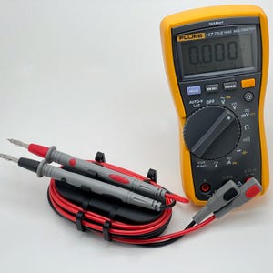 Peut inclure: Un multimètre Fluke 117 True RMS jaune avec un écran numérique affichant "0.000". Comprend des fils de test rouges et noirs et un support noir. Le multimètre a divers boutons de fonction et un cadran pour différents réglages.