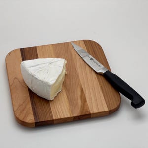 Puede incluir: Una cuña de queso brie blanco sobre una tabla de cortar de madera con un cuchillo de mango negro.
