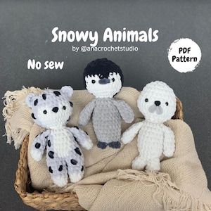 Pode incluir: Três animais de pelúcia de crochê: um pinguim, uma foca e um leopardo. Os animais são feitos com fio branco e cinza e estão sentados em uma cesta de vime forrada com um tecido bege. O texto "Snowy Animals" e "No sew" é visível na imagem.