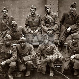 May include: A black and white historical photograph of eight soldiers in military uniforms, including long coats and peaked caps. The group is arranged in two rows, with four individuals standing in the back and four crouching in the front.
