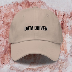Op de afbeelding: Een beige baseballpet met zwarte geborduurde tekst die "DATA DRIVEN" luidt.