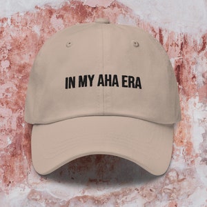 Puede incluir: Una gorra de béisbol beige con texto bordado en negro que dice "IN MY AHA ERA".