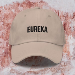 Puede incluir: Una gorra de béisbol beige con bordado negro que dice "EUREKA".