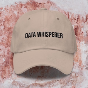 May include: A beige baseball cap with the words "DATA WHISPERER" printed in black. The cap has a curved peak and is made of a soft fabric. The background is a textured, mottled pink and white.