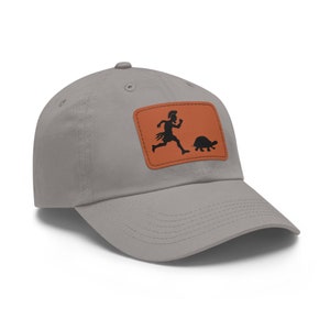 May include: A gray baseball cap with a brown leather patch featuring a silhouette of a person running after a turtle. The patch has a stitched border.