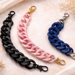 Puede incluir: Tres correas de cadena gruesas en negro, rosa y azul, cada una con un cierre de mosquetón. Las cadenas están hechas de eslabones grandes y redondeados. La cadena rosa tiene un cierre dorado, la negra tiene un cierre negro y la azul tiene un cierre plateado.