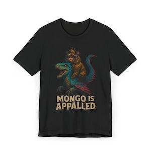 Può includere: T-shirt nera con un'illustrazione umoristica di un gatto con occhiali da sole e corona, in sella a un dinosauro. Il testo "MONGO IS APPALLED" è stampato sotto in un font vintage.