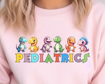 Puede incluir: Una sudadera rosa claro con una fila de dinosaurios de dibujos animados coloridos sobre la palabra "PEDIATRICS" en una fuente de arcoíris. Los dinosaurios son de varios colores, incluyendo azul, amarillo, morado, verde, naranja y rosa.
