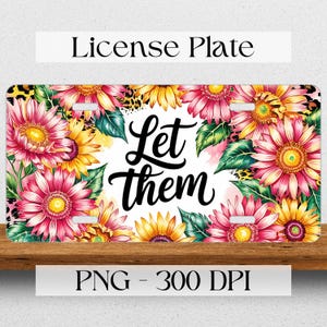 May include: A white license plate with a floral design featuring pink and yellow sunflowers. The text "Let them" is written in black cursive lettering.
